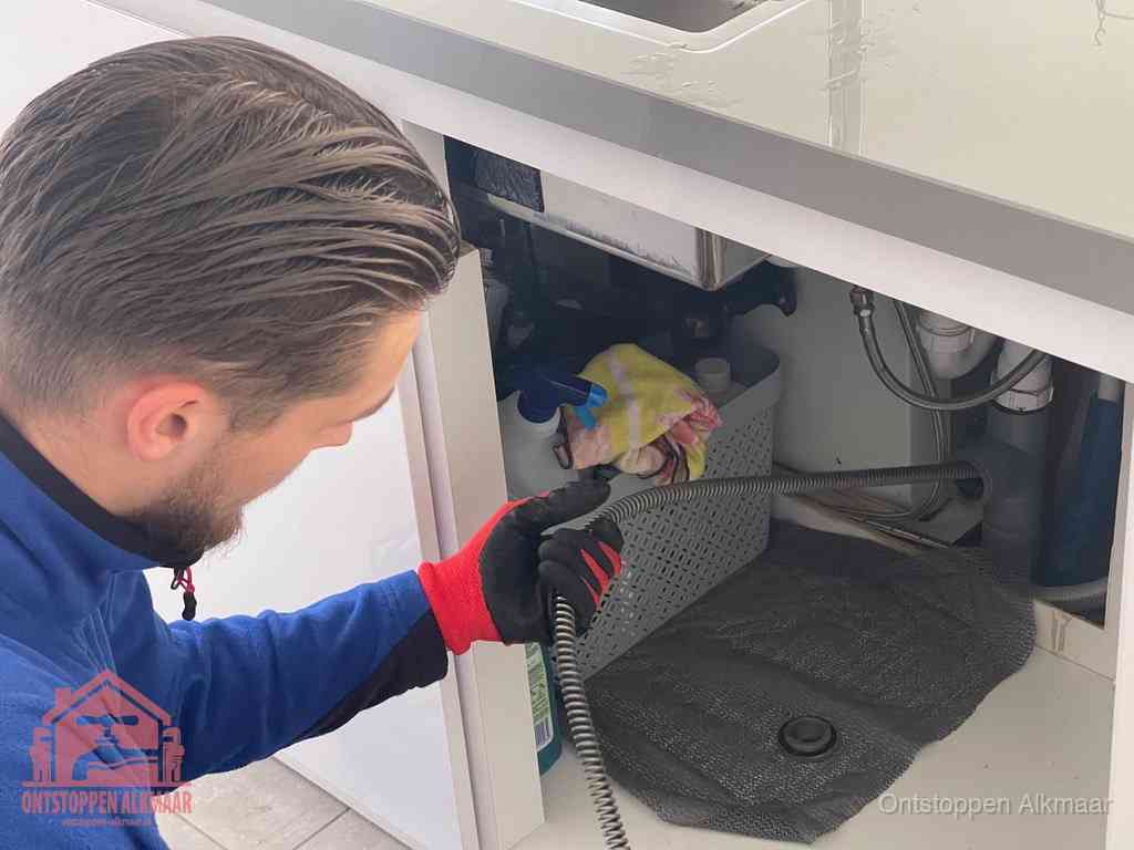 Een monteur voert een flexibele ontstoppingsveer in onder het keukenkastje, langs de sifon, om de gootsteen te ontstoppen in Alkmaar