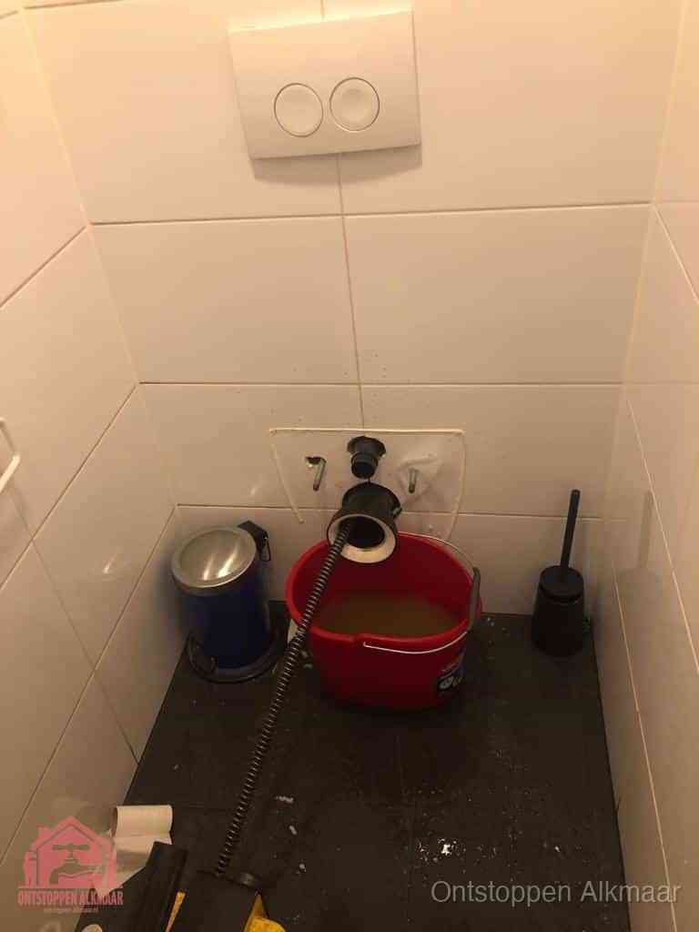Een ontstopte wand-wc met verwijderde toiletpot waar een ontstoppingsveer in de wanduitgang verdwijnt, het achterhalen waar de verstopping zit in Alkmaar