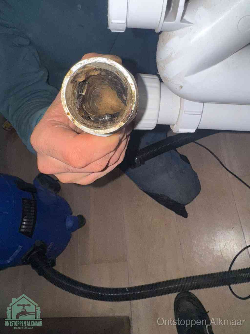 Een hand houdt een losgekoppeld PVC-afvoerbuisstuk vast dat van binnen volledig is dichtgeslibd met vet, vuil en aanslag, de meest voorkomende oorzaak van een verstopte afvoer in Alkmaar
