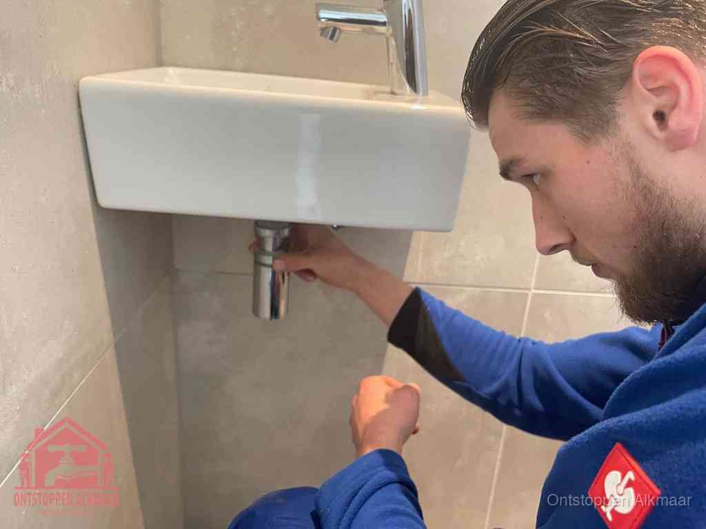 Een monteur draait met de hand de zwanenhalssifon onder een fonteinwasbak in de toiletruimte los om de bron van rioollucht in Alkmaar te vinden