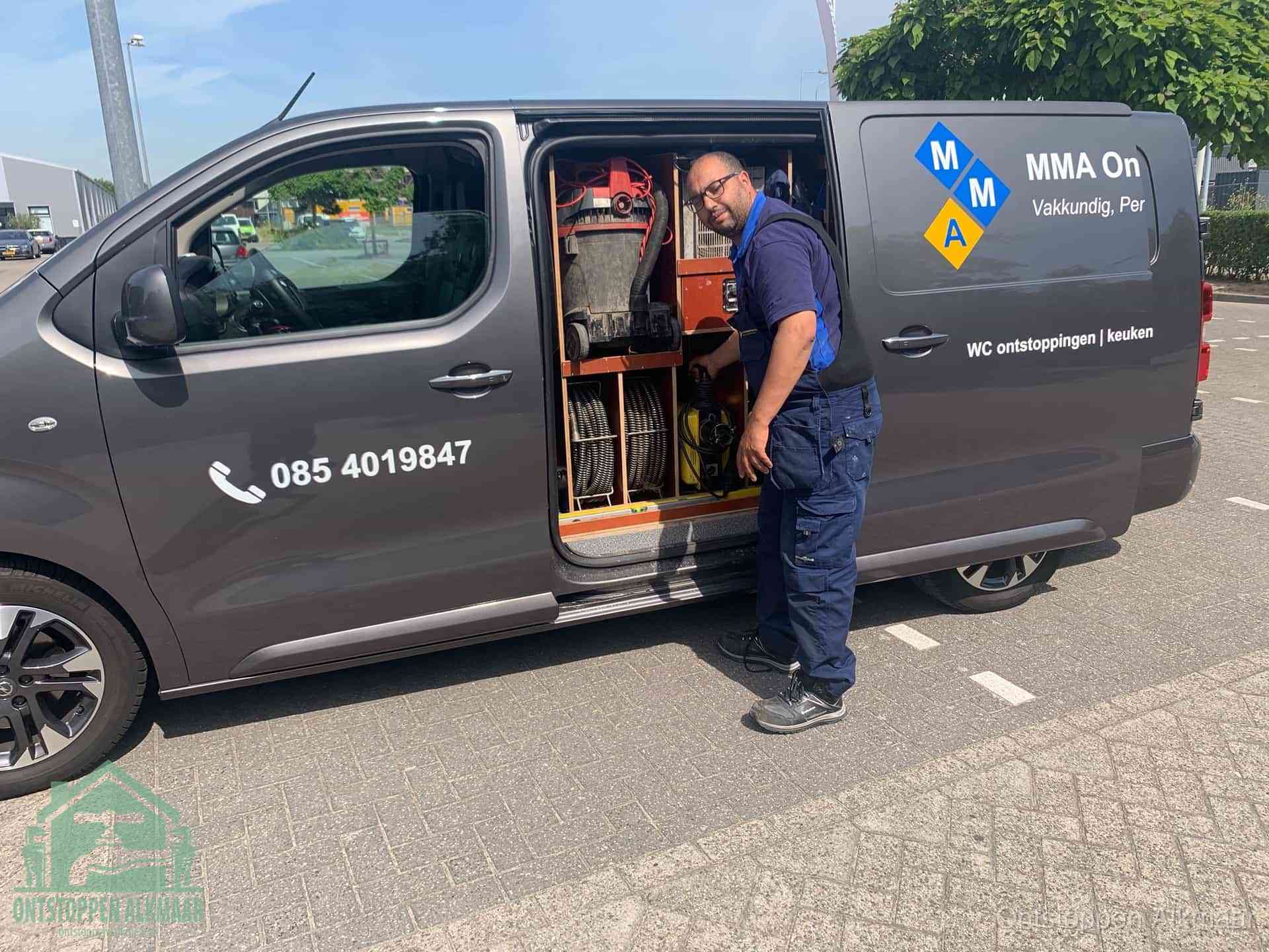 Een monteur bij de open schuifdeur van zijn bedrijfsbus op een parkeerplaats, klaar om de oorzaak van terugkerende afvoerverstoppingen aan te pakken in Alkmaar