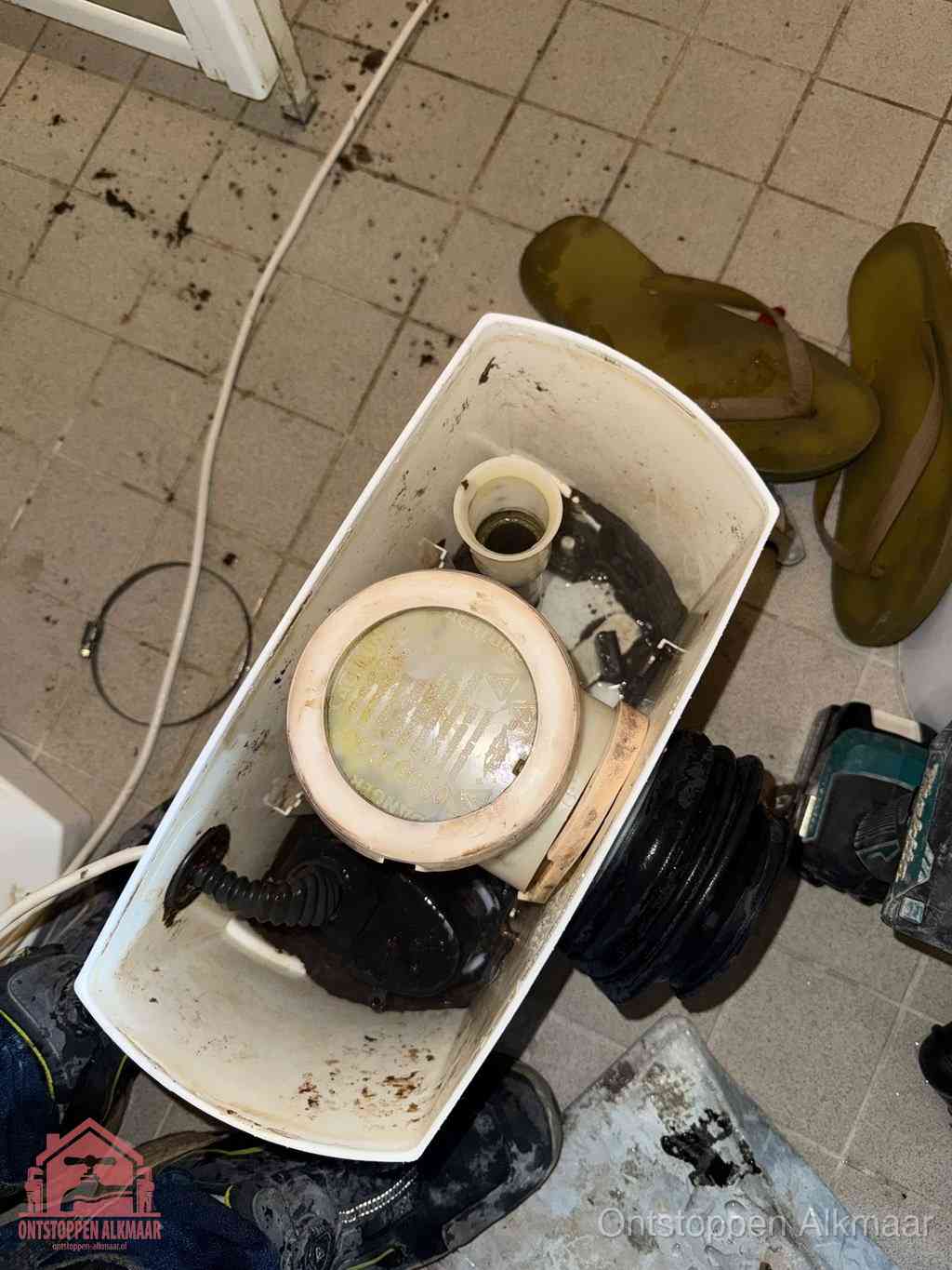 Een gedemonteerde toiletpot op de vloer met ontstoppingsveer en gereedschap ernaast, het verwijderen van een vastzittend voorwerp uit de wc in Alkmaar
