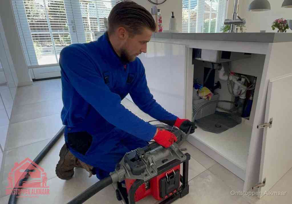 Een monteur voert een professionele elektrische ontstoppingsmachine in de afvoer onder de spoelbak tijdens het gootsteen ontstoppen in Alkmaar